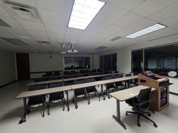 Large hybrid learning classroom with rows of desks, a ceiling-mounted projector, and integrated instructor station by The Brass Effect.